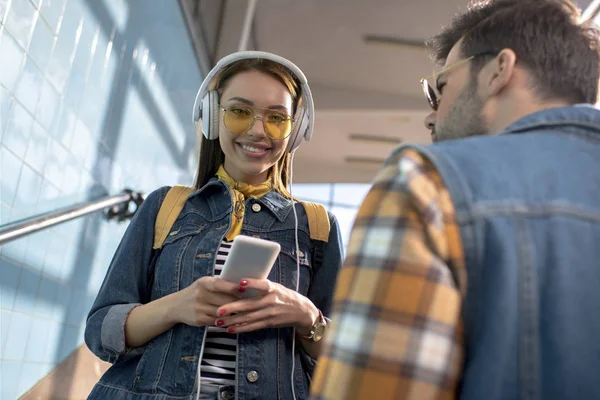 Turista femenina con estilo sonriente en gafas de sol con auriculares con teléfono inteligente y novio de pie cerca en el metro - foto de stock