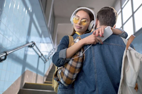 Jeune touriste féminine élégante dans des lunettes de soleil avec écouteurs et smartphone étreignant petit ami au métro — Photo de stock