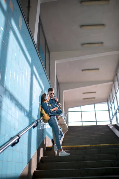 Low angle view of stylish female tourist with headphones showing smartphone to boyfriend in sunglasses at subway — Stock Photo