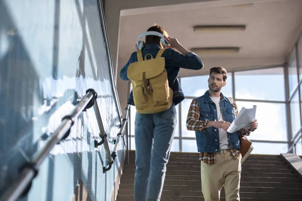 Vista trasera de la mujer con estilo quitando auriculares para ayudar a turista masculino con mapa en las manos en el metro - foto de stock