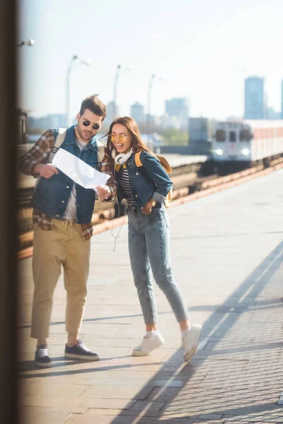Elegante pareja de viajeros con mochilas en gafas de sol mirando el mapa en la estación de metro al aire libre - foto de stock