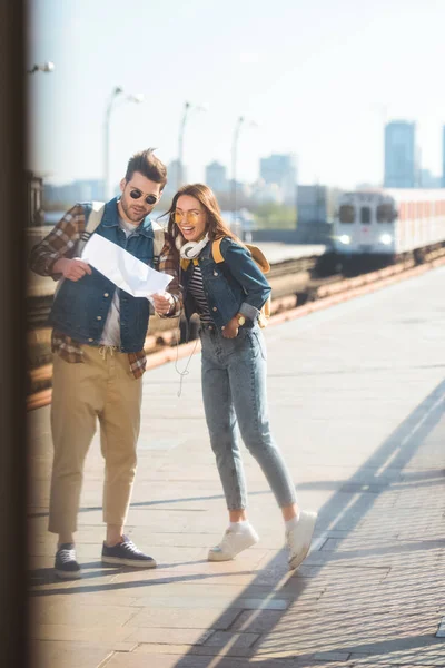 Stylish couple of travelers with backpacks in sunglasses looking at map at outdoor subway station — Stock Photo