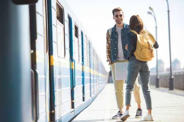 Visão traseira de turista elegante feminino com mochila conversando com namorado sorridente em óculos de sol na estação de metrô ao ar livre — Fotografia de Stock