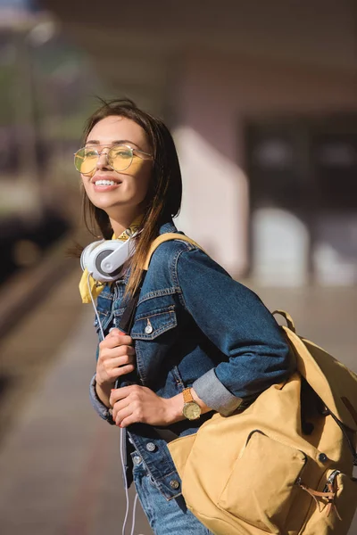 Mulher elegante em óculos de sol com mochila e fones de ouvido em pé na estação de metro ao ar livre — Fotografia de Stock