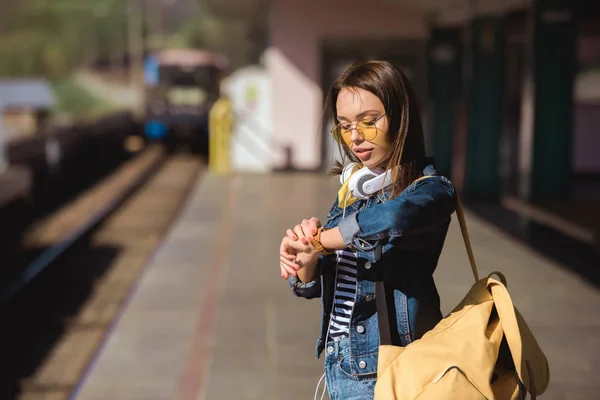 Стильная женщина в солнечных очках в наушниках и рюкзаке, смотрящая на наручные часы — стоковое фото