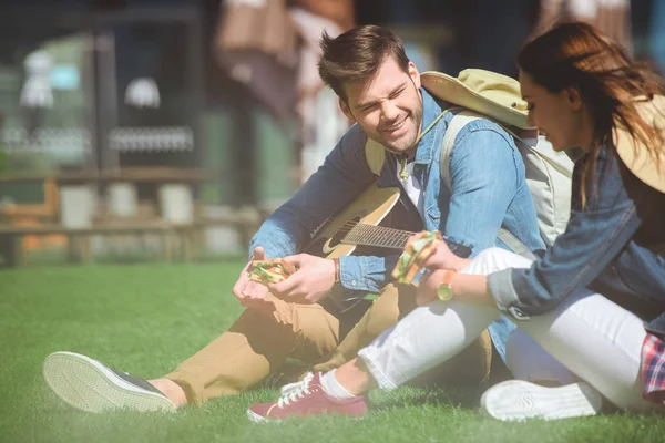 Sorrindo casal de viajantes com guitarra comendo sanduíches e sentado na grama — Fotografia de Stock