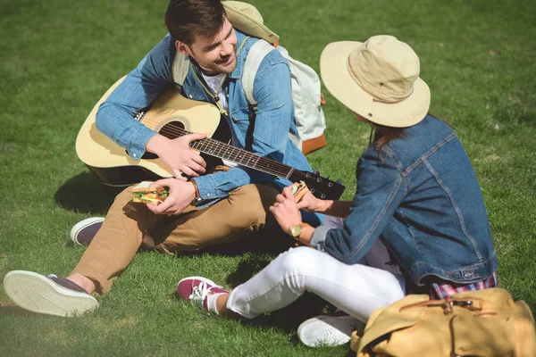 Couple of stylish tourists with backpacks and guitar sitting on grass and eating sandwiches — Stock Photo