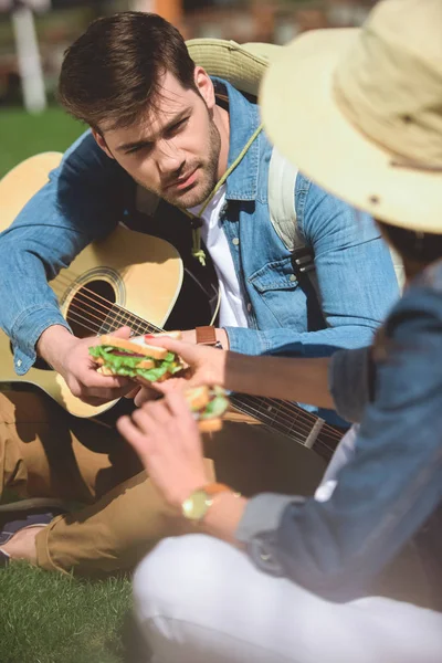 Rear view of stylish female tourist giving sandwich to boyfriend with guitar — Stock Photo