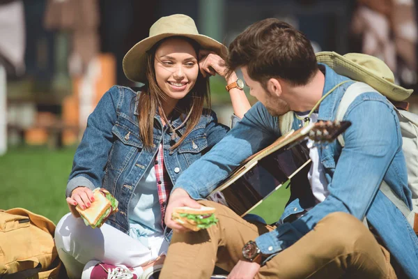 Stylish travelers in hats with backpacks and guitar holding sandwiches — Stock Photo