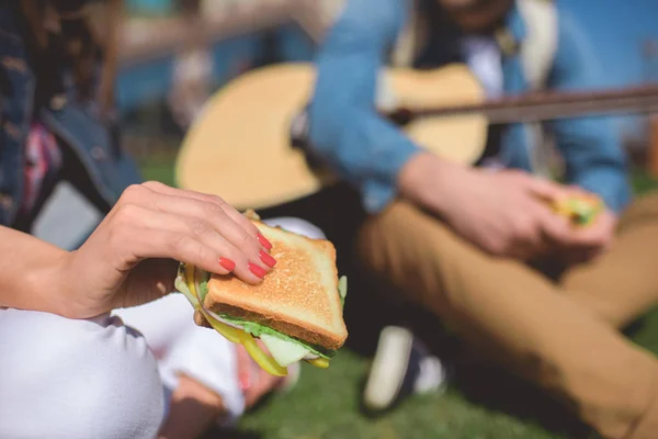 Close-up tiro de sanduíche na mão feminina e homem sentado com guitarra acústica — Fotografia de Stock