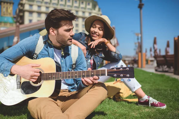 Turista masculino jogando na guitarra e namorada olhando para ele na grama — Fotografia de Stock