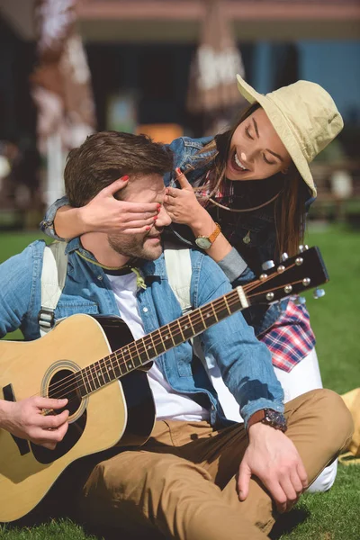 Sonriente viajero femenino en sombrero cubriendo los ojos de novio con guitarra por detrás - foto de stock