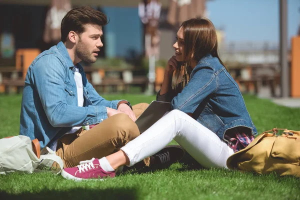 Side view of stylish couple of travelers with backpacks using laptop on grass — Stock Photo