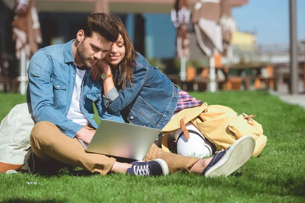 Jovem casal elegante de viajantes com mochilas usando laptop na grama — Fotografia de Stock