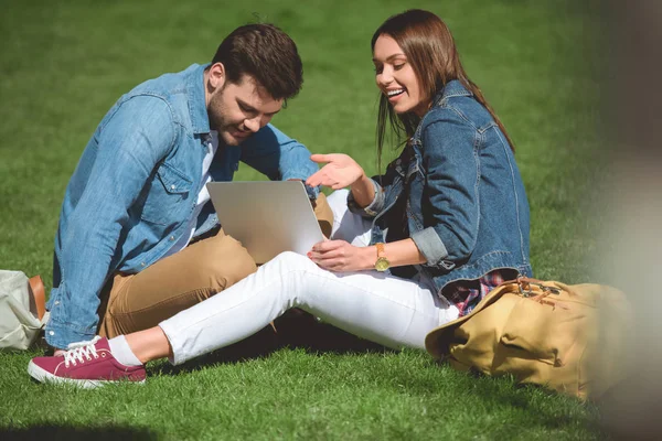 Couple of stylish young tourists with backpacks and laptop sitting on grass — Stock Photo