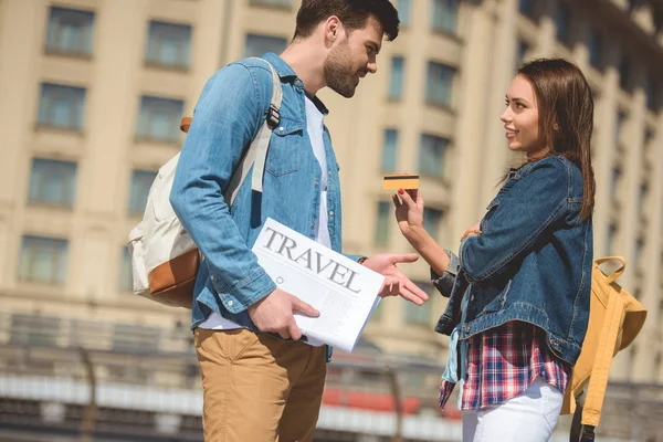 Sonriente turista mujer dando tarjeta de crédito a novio con periódico de viaje en la mano - foto de stock