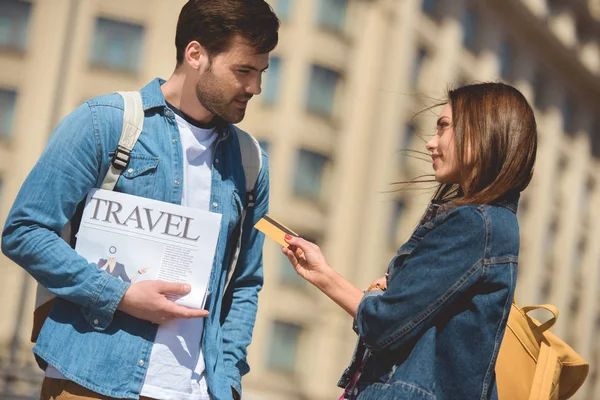 Stylish female tourist giving credit card to boyfriend with travel newspaper in hand — Stock Photo