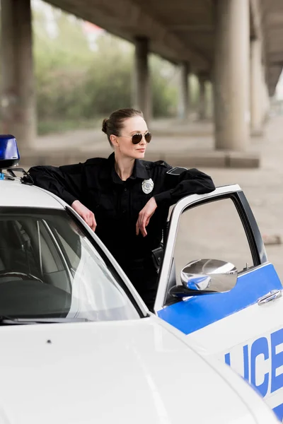 Atractiva mujer oficial de policía de pie cerca de coche patrulla - foto de stock