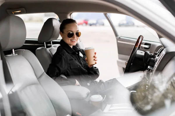 Smiling policewoman with paper cup of coffee sitting in car and looking at camera — Stock Photo