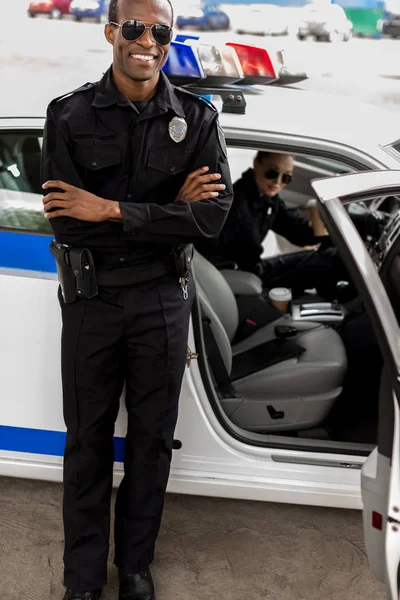 Apuesto joven policía con los brazos cruzados de pie delante de coche de policía - foto de stock