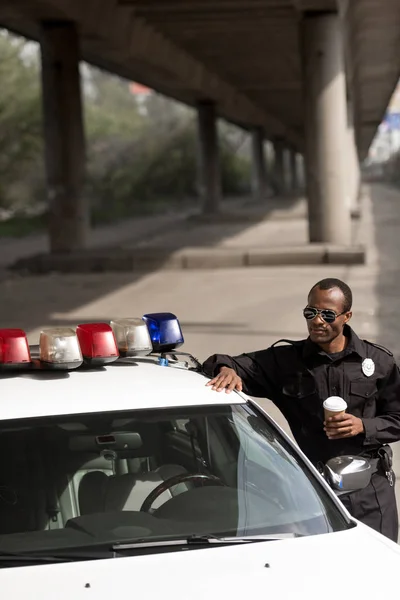 Oficial de policía con café para ir apoyándose en coche de policía en la calle — Stock Photo