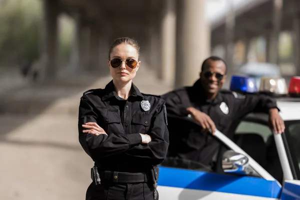 Jeune policière sérieuse debout avec les bras croisés tandis que son partenaire debout près de la voiture et souriant flou sur fond — Photo de stock