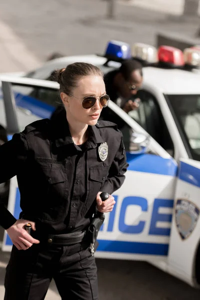 Young serious policewoman standing arms akimbo and looking away while her partner standing near car blurred on background — Stock Photo