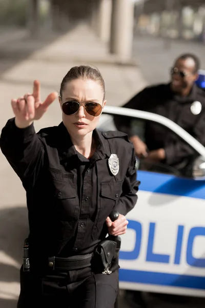 Молода серйозна поліцейська вказує десь, поки її партнер стоїть біля машини розмитий на фоні — стокове фото