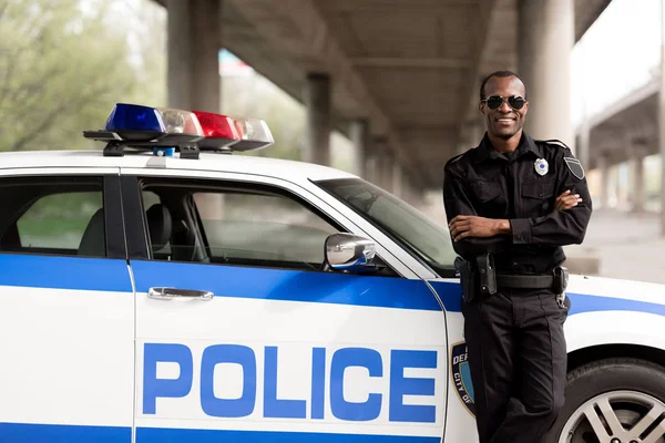 Handsome african american police officer with crossed arms leaning back on car and looking at camera — Stock Photo