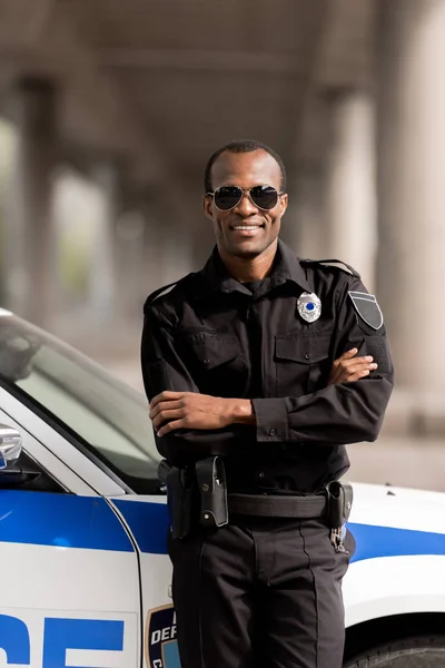 Lächelnder afrikanisch-amerikanischer Polizist mit verschränkten Armen, der sich auf sein Auto zurücklehnt und in die Kamera blickt — Stockfoto