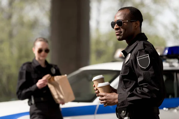 Jeunes policiers avec café pour aller et sac en papier avec déjeuner ayant pause — Photo de stock