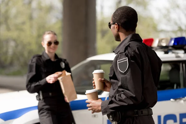 Jóvenes agentes de policía con café para llevar y bolsa de papel con almuerzo teniendo descanso al lado del coche - foto de stock