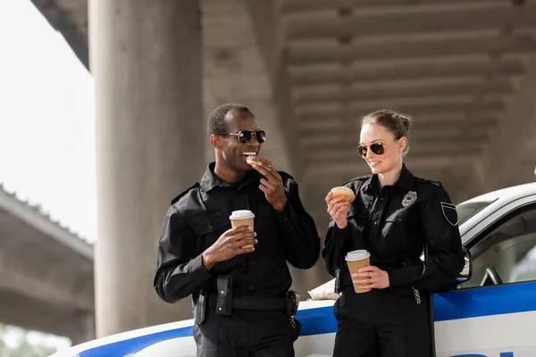 Jeunes policiers avec café et beignets debout à côté de la voiture sous le pont — Photo de stock