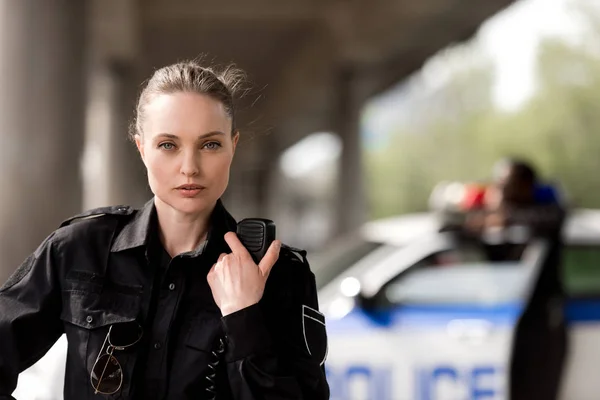 Policewoman using walkie-talkie and looking at camera with blurred partner near car on background — Stock Photo
