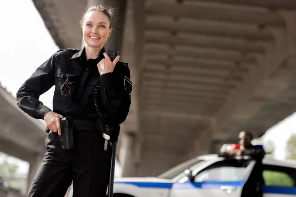 Счастливая привлекательная женщина-полицейский, использующая рацию с размытым партнером рядом с машиной на заднем плане — стоковое фото