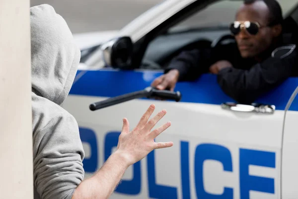 Police officer peering out car window and pointing at hooded man with police bat — Stock Photo