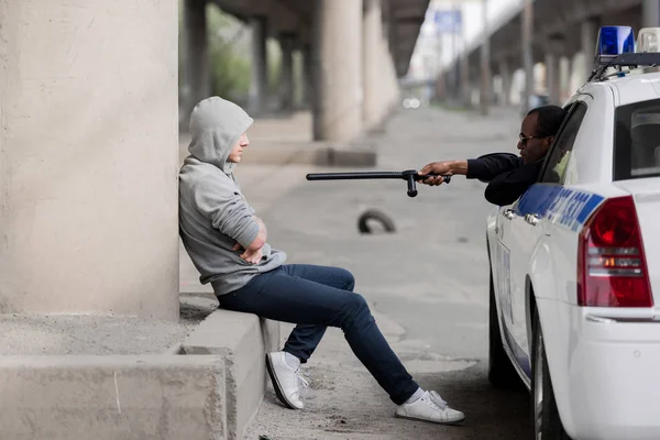 Police officer pointing at hooded man with police bat while he sitting on street — Stock Photo