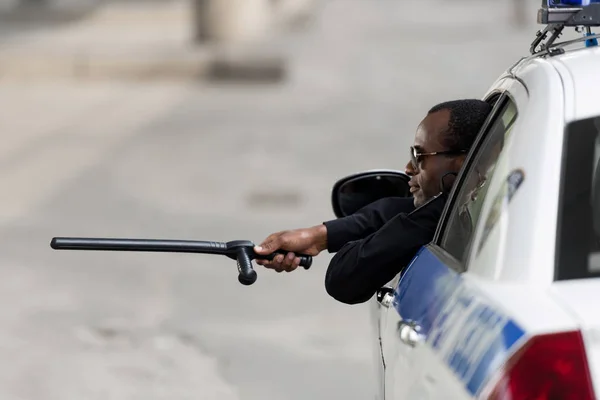 African american police officer peering out of window of police car with bat — Stock Photo