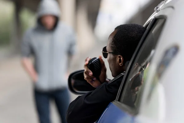 Policía mirando a un joven encapuchado en la calle por la ventana y hablando por radio - foto de stock