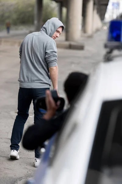 Policeman looking on hooded young man walking away on street out of window — Stock Photo