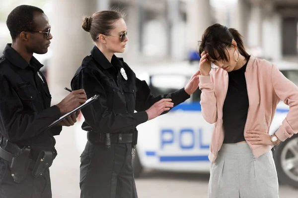 African american policeman writing in clipboard and policewoman cheering up upset young woman — Stock Photo