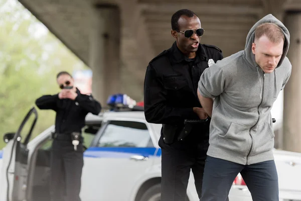 Irritado policial afro-americano prendendo jovem enquanto parceiro feminino cobrindo-o com arma — Fotografia de Stock