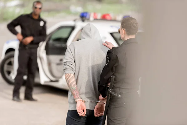 Вид ззаду поліцейської, яка заарештовує злочинця в наручниках, а партнер стоїть за автомобілем — стокове фото