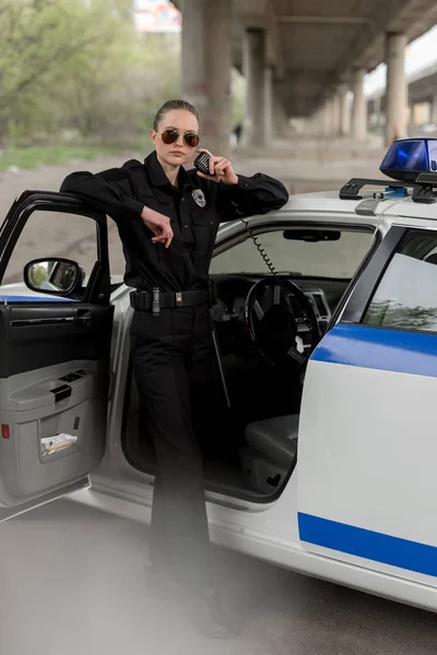 Policewoman in sunglasses standing near car and talking on portable radio — Stock Photo