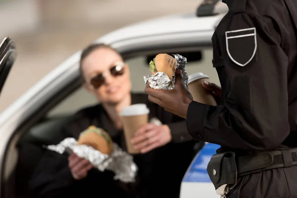 Imagen recortada de policía y policía con tazas de café y hamburguesas - foto de stock