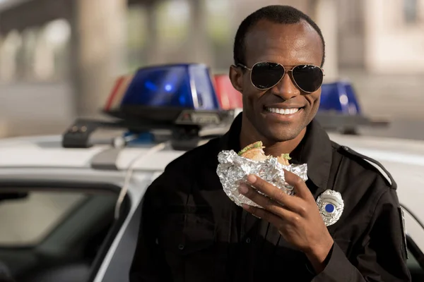 Smiling african american policeman in sunglasses with burger in hand near car — Stock Photo