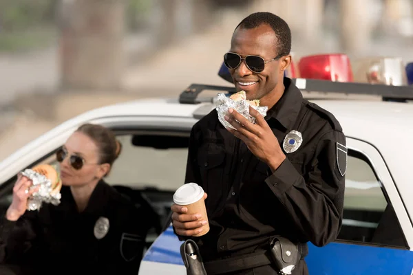 African american policeman with paper cup of coffee eating burger while his female partner eating burger in car — Stock Photo