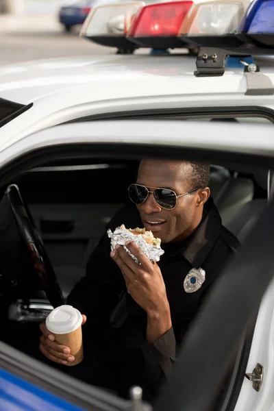 Young african american policeman with coffee cup sitting in car and eating burger — Stock Photo