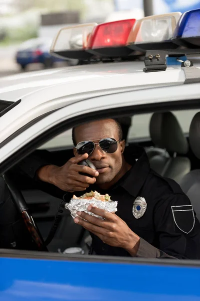 Policía afroamericano sosteniendo hamburguesa y hablando por radio portátil en coche — Stock Photo