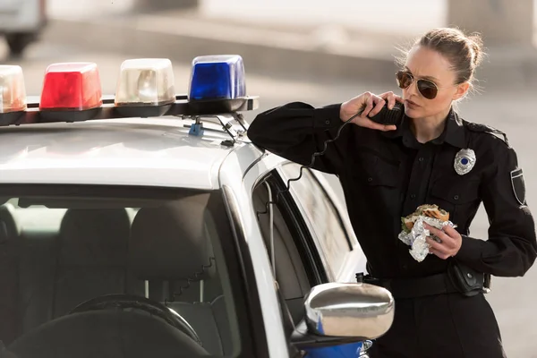 Serious policewoman with burger in hand talking on radio set — Stock Photo